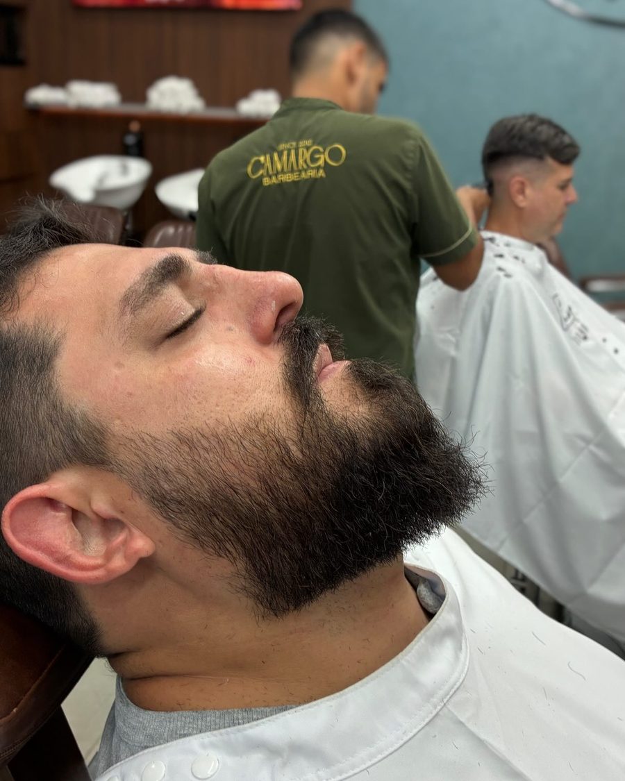 Cliente com a barba sendo feita, e um barbeiro atendendo ao fundo