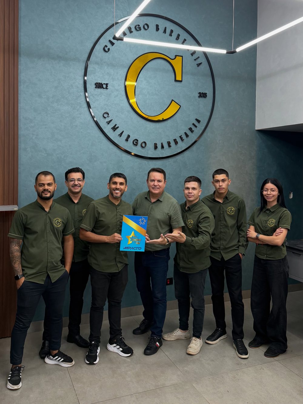 Equipe da Camargo Barbearia de uniforme verde, diante do letreiro da parede